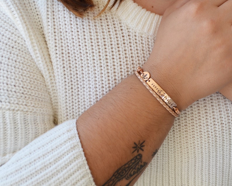 K&ouml;nnte beinhalten: Ein ros&eacute;goldenes Armband mit einem geflochtenen Lederband und einem rechteckigen Anh&auml;nger mit dem eingravierten Wort "DUFF". Das Armband hat auch einen kleinen runden Anh&auml;nger mit einem Kompass-Design. Das Armband wird an einem Handgelenk getragen.