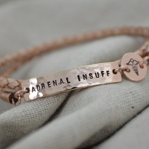 K&ouml;nnte beinhalten: Ein ros&eacute;goldenes medizinische Armband mit den Worten "ADRENAL INSUFF" auf einer geh&auml;mmerten Metallplatte. Das Armband hat ein geflochtenes Lederband und einen kleinen medizinischen Symbolanh&auml;nger.