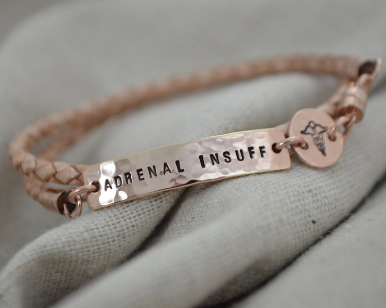 K&ouml;nnte beinhalten: Ein ros&eacute;goldenes medizinische Armband mit den Worten "ADRENAL INSUFF" auf einer geh&auml;mmerten Metallplatte. Das Armband hat ein geflochtenes Lederband und einen kleinen medizinischen Symbolanh&auml;nger.