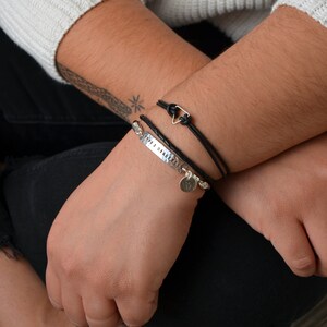 May include: A person wearing a white sweater and black pants, with two bracelets on the left wrist. One bracelet is silver with the text "2012 08 04" and charms. The other is a black braided leather bracelet with a silver triangle clasp.