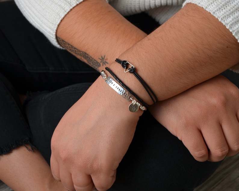 May include: A person wearing a white sweater and black pants, with two bracelets on the left wrist. One bracelet is silver with the text "2012 08 04" and charms. The other is a black braided leather bracelet with a silver triangle clasp.