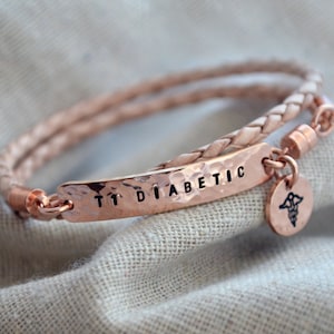 May include: A rose gold medical alert bracelet with a braided leather band. The hammered metal plate reads "T1 DIABETIC" in black letters. A small round charm with a medical symbol hangs from the bracelet.