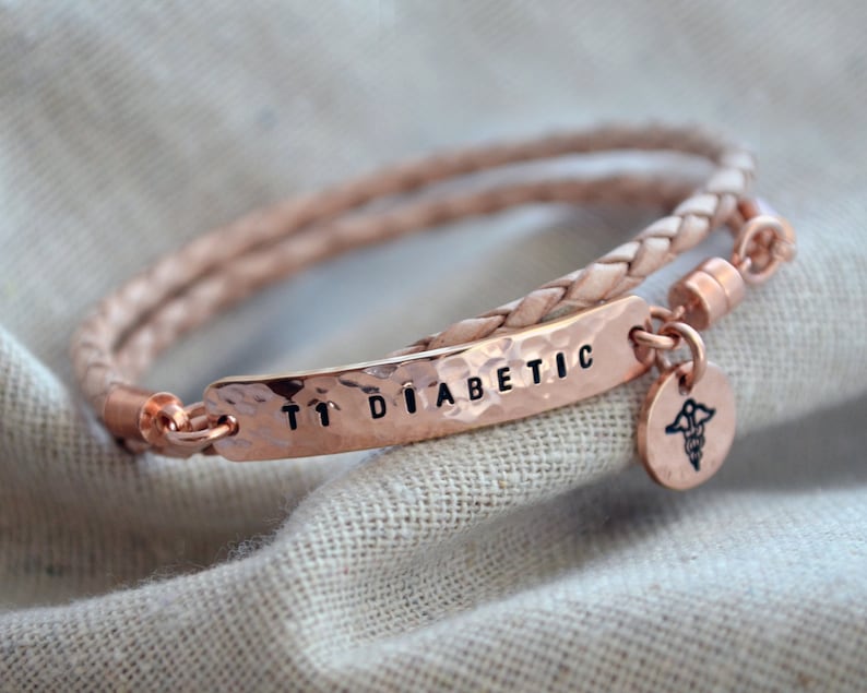 May include: A rose gold medical alert bracelet with a braided leather band. The hammered metal plate reads "T1 DIABETIC" in black letters. A small round charm with a medical symbol hangs from the bracelet.