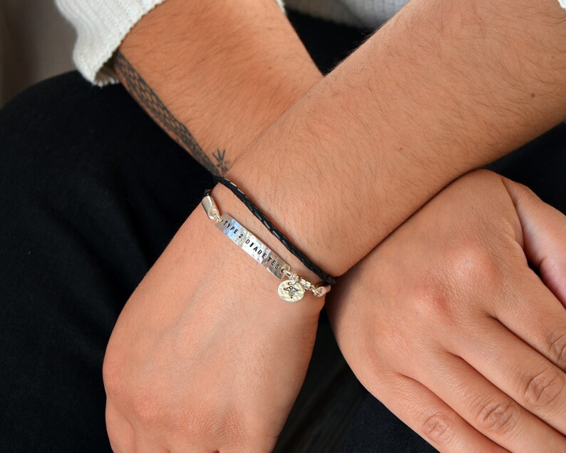 May include: Two bracelets are shown. One is a black braided leather bracelet with a silver charm. The other is a silver bracelet with the words "TYPE 2 DIABETES" stamped on it. The bracelets are worn on a person's wrist.