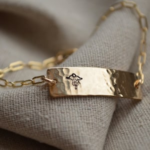 May include: A gold-colored medical ID bracelet with a hammered texture. The rectangular plate features a black caduceus symbol and is connected to a chain-link bracelet. The bracelet is displayed on a neutral-colored fabric.