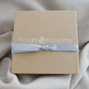 May include: A square, light brown gift box with a gray ribbon tied around it. The box has the words "thought blossoms" printed in white on the top. The box is sitting on a beige fabric background.