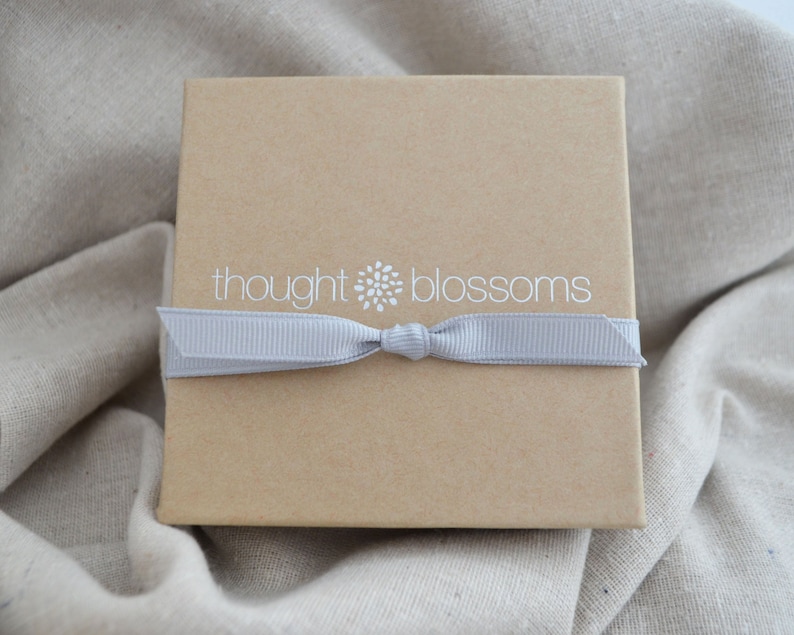 May include: A square, light brown gift box with a gray ribbon tied around it. The box has the words "thought blossoms" printed in white on the top. The box is sitting on a beige fabric background.
