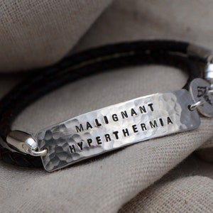 May include: A medical alert bracelet with a dark brown braided leather band and a hammered silver plate. The plate is stamped with the words "MALIGNANT HYPERTHERMIA". A small silver charm with a medical symbol hangs from the clasp.