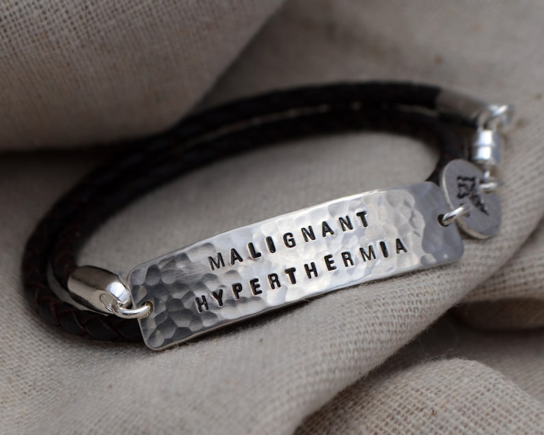 May include: A medical alert bracelet with a dark brown braided leather band and a hammered silver plate. The plate is stamped with the words "MALIGNANT HYPERTHERMIA". A small silver charm with a medical symbol hangs from the clasp.
