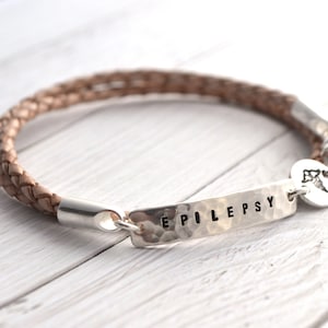 K&ouml;nnte beinhalten: Ein geflochtenes Lederarmband in Beige mit einem silbernen Balken, auf dem das Wort "EPILEPSY" steht. Ein kleiner Silberanh&auml;nger mit einem medizinischen Symbol ist angebracht.