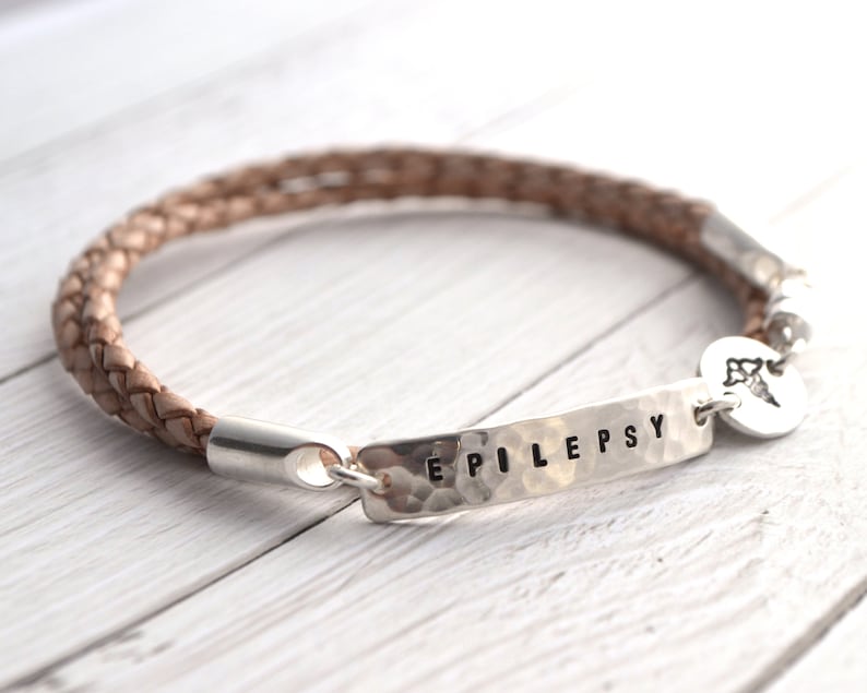 K&ouml;nnte beinhalten: Ein geflochtenes Lederarmband in Beige mit einem silbernen Balken, auf dem das Wort "EPILEPSY" steht. Ein kleiner Silberanh&auml;nger mit einem medizinischen Symbol ist angebracht.
