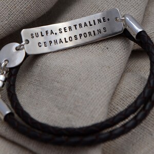 May include: A silver bracelet with a dark brown braided leather band. The rectangular silver tag is stamped with the words: SULFA, SERTRALINE, CEPHALOSPORINS. The bracelet has a magnetic clasp.
