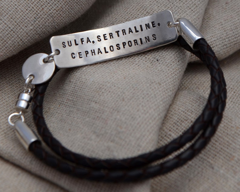 May include: A silver bracelet with a dark brown braided leather band. The rectangular silver tag is stamped with the words: SULFA, SERTRALINE, CEPHALOSPORINS. The bracelet has a magnetic clasp.