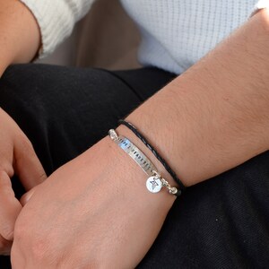 May include: A silver bracelet with the text "TYPE 2 DIABETES" and a medical symbol charm. The bracelet is paired with a black braided leather bracelet. The bracelets are worn on a person's wrist.