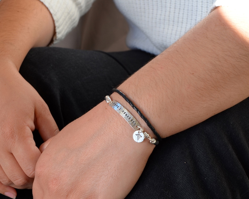 May include: A silver bracelet with the text "TYPE 2 DIABETES" and a medical symbol charm. The bracelet is paired with a black braided leather bracelet. The bracelets are worn on a person's wrist.