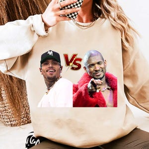May include: Beige sweatshirt featuring a graphic of Chris Brown and Usher with a "Vs" logo. Chris Brown is wearing a black cap and glasses. Usher is wearing a red fur coat and pointing. The sweatshirt is a neutral color.