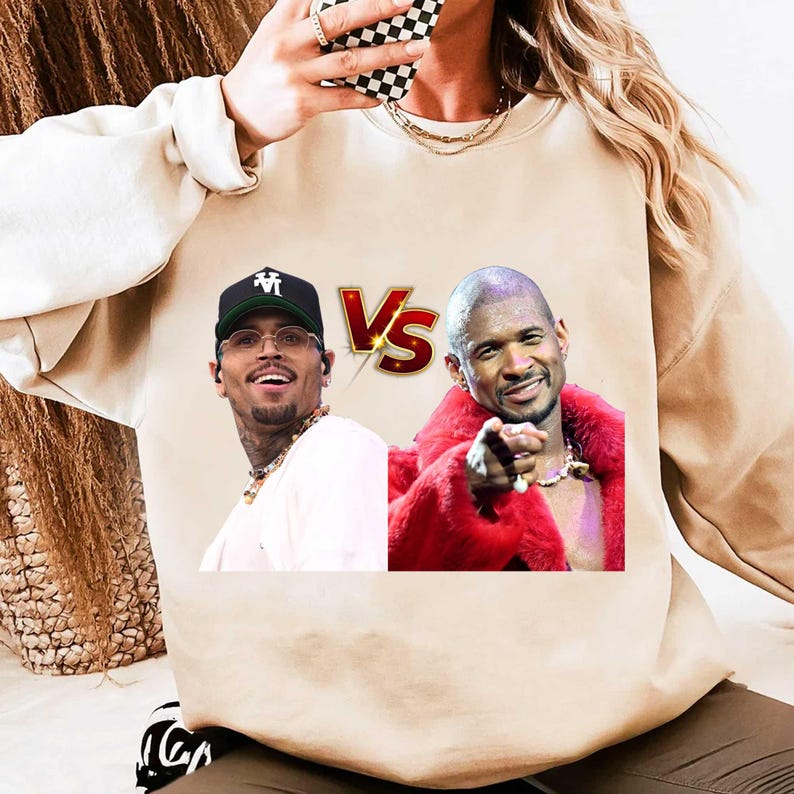 May include: Beige sweatshirt featuring a graphic of Chris Brown and Usher with a "Vs" logo. Chris Brown is wearing a black cap and glasses. Usher is wearing a red fur coat and pointing. The sweatshirt is a neutral color.