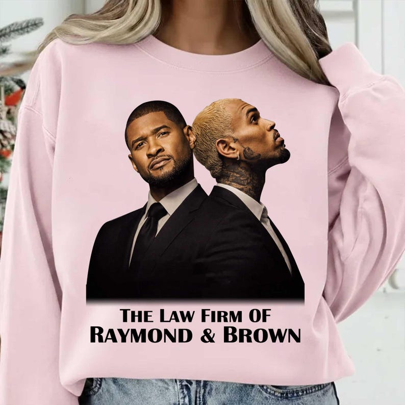 May include: A light pink sweatshirt featuring a graphic of two men in black suits. The text "THE LAW FIRM OF RAYMOND & BROWN" is printed in black at the bottom of the graphic. The sweatshirt has long sleeves.