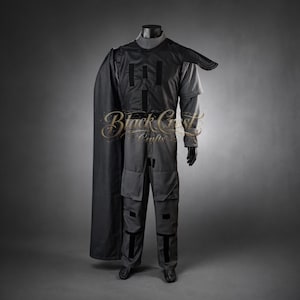 Pode incluir: Um traje cinza escuro com capa, exibido em um manequim. O traje inclui um top de gola alta, calças com bolsos e uma capa. O traje tem detalhes em preto e provavelmente é para cosplay ou uso teatral. O texto "Black Crest Crafts" é visível.
