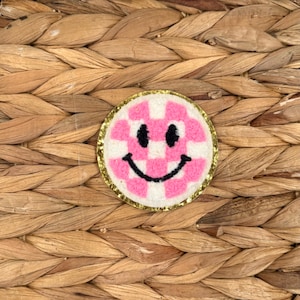 May include: A round patch with a pink and white checkered smiley face design. The patch has a gold glitter border and a black stitched smile and eyes. The patch is on a woven background.