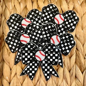 May include: A collection of baseball-themed hair bows. Each bow features a black and white chequered pattern with a baseball in the centre. The baseballs have red stitching. The bows are arranged on a woven, textured surface.
