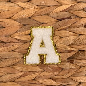 May include: A white felt letter "A" with a gold glitter border. The letter is set against a woven, natural-toned background. This decorative patch is suitable for various craft projects.