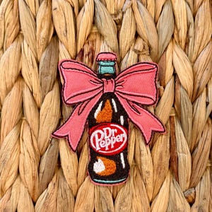 Puede incluir: Parche bordado con una botella de Dr. Pepper con un lazo rosa. El parche tiene un contorno negro e incluye el logotipo de Dr. Pepper en un círculo blanco con texto rojo. El fondo es una textura tejida.