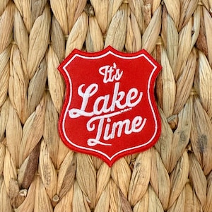 Puede incluir: Un parche rojo en forma de escudo con texto bordado en blanco que dice "It's Lake Time". El parche tiene un borde blanco y está sobre un fondo tejido de color marrón claro. El parche es probablemente para ropa o accesorios.