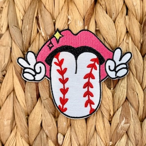 Puede incluir: Un parche con temática de béisbol que presenta una boca rosa con un contorno negro y una estrella. La lengua está diseñada como una pelota de béisbol con costuras rojas. Manos blancas con signos de paz están a cada lado de la boca. El parche está sobre un fondo tejido.