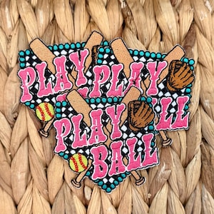 May include: A softball-themed patch with the words "PLAY BALL" in pink, outlined in white, and set against a black and teal background. The design includes baseball bats, gloves, and softballs.