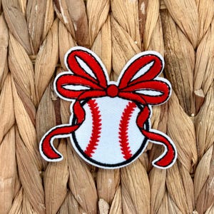 Könnte beinhalten: Gestickter Aufnäher mit einem Baseball mit roten Nähten und einer roten Schleife. Der Baseball ist weiß mit roten Nähten, und die Schleife ist rot mit weißem Rand. Der Aufnäher hat einen schwarzen Rand.