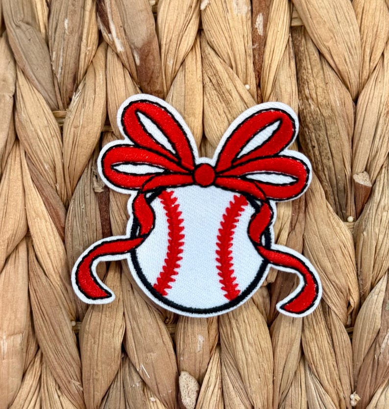 May include: Embroidered patch featuring a baseball with red stitching and a red bow. The baseball is white with red stitching, and the bow is red with a white outline. The patch has a black outline.