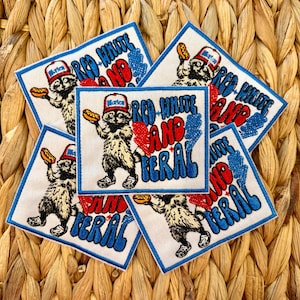 May include: Embroidered patches with a raccoon holding a hot dog, wearing a hat that says "Merica". The patches have the words "Red White and Feral" in red and blue. The patches are white with a blue border and are arranged on a woven background.