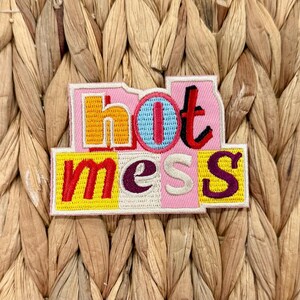 May include: Embroidered patch with the words "hot mess" in a colorful, blocky design. The patch features a pink background with yellow, blue, red, and purple lettering. The patch is set against a woven, natural fiber background.