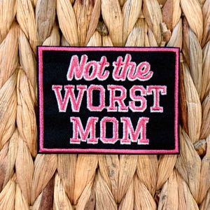 May include: A rectangular black patch with the words "Not the WORST MOM" embroidered in pink and white. The patch has a pink border and is set against a woven background.