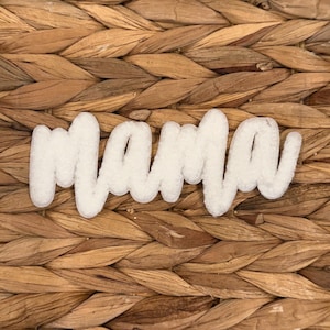 Może przedstawiać: Białe filcowe słowo "Mama" w kursywie, umieszczone na tkanym, brązowym tle. Słowo jest punktem centralnym, o miękkim, teksturowanym wyglądzie. Tło to naturalny, teksturowany splot.
