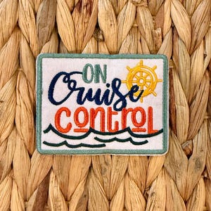 Puede incluir: Parche bordado con las palabras "ON Cruise Control" en azul marino y naranja, con un timón amarillo y olas verdes. El parche tiene un fondo blanco y un borde verde.