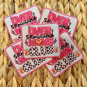 May include: A collection of square patches with the words "OVER STIMULATED MOMS CLUB" in pink and black lettering. The patches have a light gray background and a pink border. The design includes peace signs, a melting smiley face, and a checkered pattern.