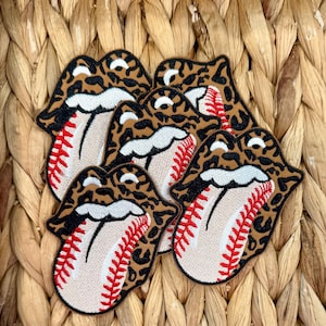 Puede incluir: Parches bordados con forma de labios con diseño de béisbol. Los labios tienen un estampado de leopardo, con una pelota de béisbol beige y costuras rojas. Los parches están colocados sobre un fondo tejido.