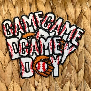 May include: Baseball-themed patches with the words "GAME DAY" in a bold, stitched font. The patches feature a baseball and glove design. The patches are white, red, and black. The patches are on a woven background.