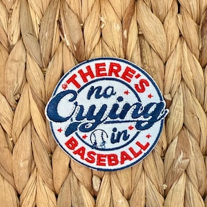 Puede incluir: Parche bordado circular con el texto "THERE'S no CRYING in BASEBALL" en rojo y azul marino. El parche presenta un gráfico de béisbol. El fondo es un material tejido de color marrón claro.