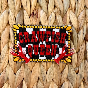 May include: A rectangular patch with the words "CRAWFISH QUEEN" in red, outlined in black, on a black and white checkered background. The patch has a yellow border with red crawfish on each side.