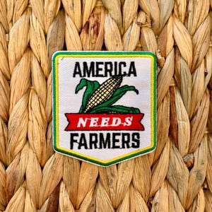 May include: A rectangular patch with a white background, green and yellow border, and the words "AMERICA NEEDS FARMERS" in black. A green corn stalk with a golden cob is in the center. The patch is set against a woven, light brown background.