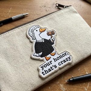 May include: A canvas pencil case with a zipper closure, featuring an embroidered patch of a goose in a judge's robe holding a gavel. The patch includes the text "your honor, that's crazy."