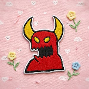 May include: An embroidered patch featuring a red monster with yellow horns and yellow eyes, outlined in black and white. The monster has a jagged black mouth and is positioned on a light pink fabric background decorated with small embroidered flowers.