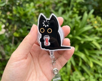 May include: A black cat badge reel featuring a white loading icon above its head and large yellow eyes. The cat is holding a silver Diet Coke can with red text. The badge reel has a clear plastic strap and a metal snap button for attachment.