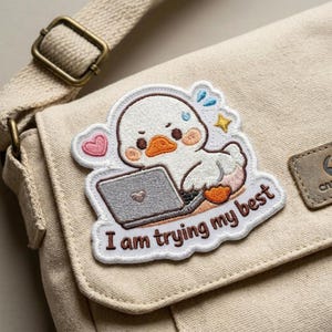 May include: Embroidered patch featuring a cute white duck working on a laptop, with the text "I am trying my best." The patch is attached to a beige canvas bag with a brown strap and buckle.