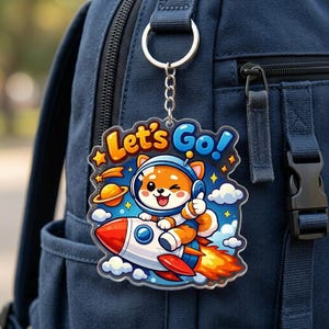 May include: A clear acrylic keychain featuring a cartoon Shiba Inu dog in a space suit riding a rocket ship. The keychain has the words "Let's Go!" in orange and blue. The keychain is attached to a silver metal ring and chain.