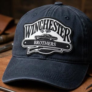 May include: Navy blue baseball cap featuring a patch with the words "Winchester Brothers" in white lettering. The patch includes a black car graphic, a rifle, and the phrases "Jerk" and "Bitch!" in a speech bubble.