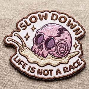May include: Embroidered patch featuring a pink skull with purple eye sockets and a snail body. The patch has the text "SLOW DOWN" and "LIFE IS NOT A RACE" in brown lettering. The patch is outlined in brown and white.